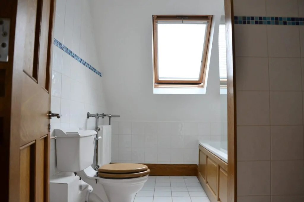 Bathroom with white tiles, a toilet, bathtub with wooden paneling, and a skylight window above.