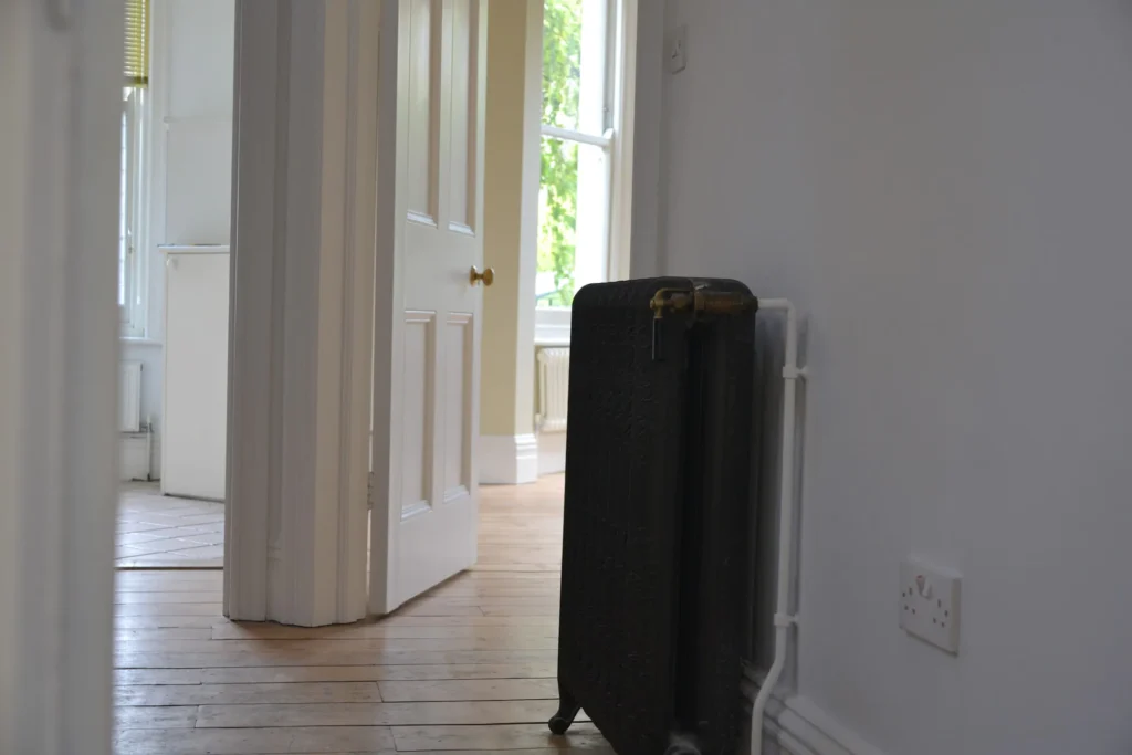 A hallway in a house with wooden floors and an open door. A radiator is visible on the right wall, and sunlight is coming through the windows in the background.