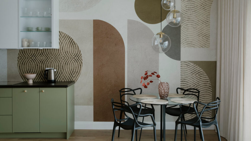 A modern dining area with a glass table, four black chairs, green cabinets, and a geometric-patterned accent wall. Three glass pendant lights hang above the table.