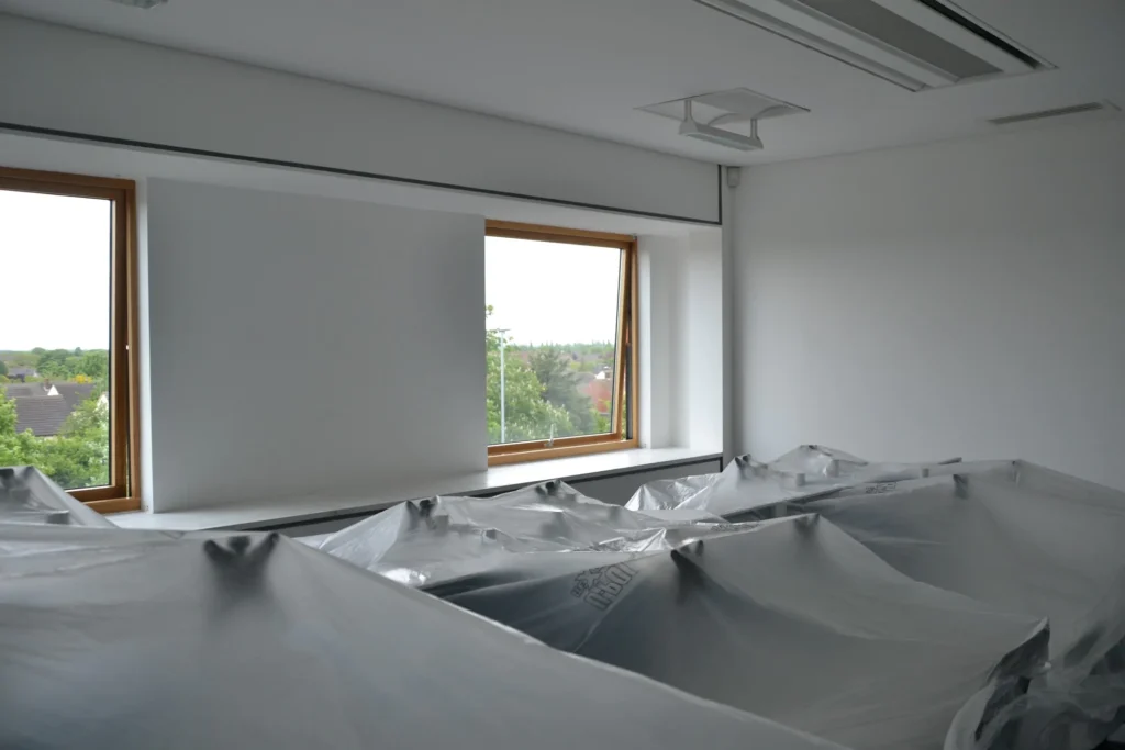 A room with covered furniture, featuring two large windows with views of trees and houses