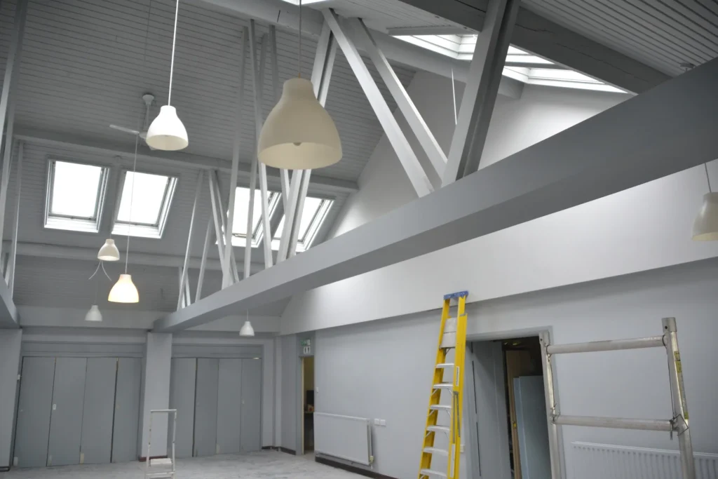 A modern room with high ceilings, large windows, pendant lights, a yellow ladder, and scaffolding. Walls and ceiling are painted gray.