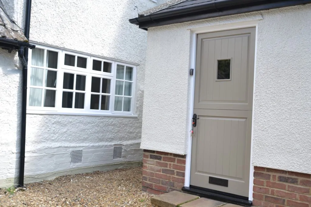 A beige wooden door in a white brick wall next to a multi-pane window with gravel ground