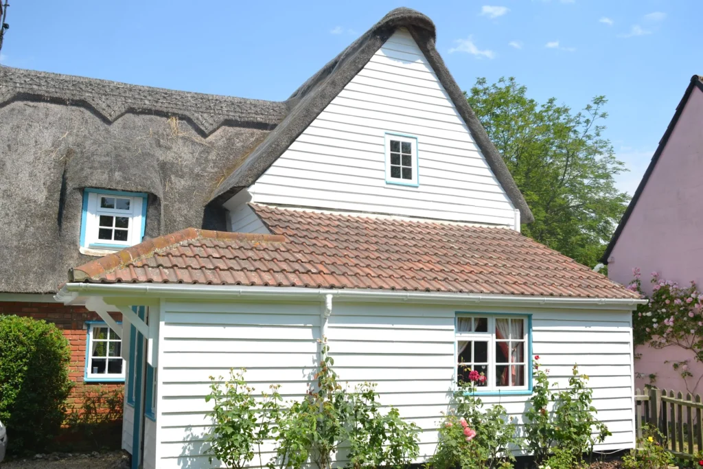 A white cottage with a thatched roof surrounded by greenery and roses in bloom