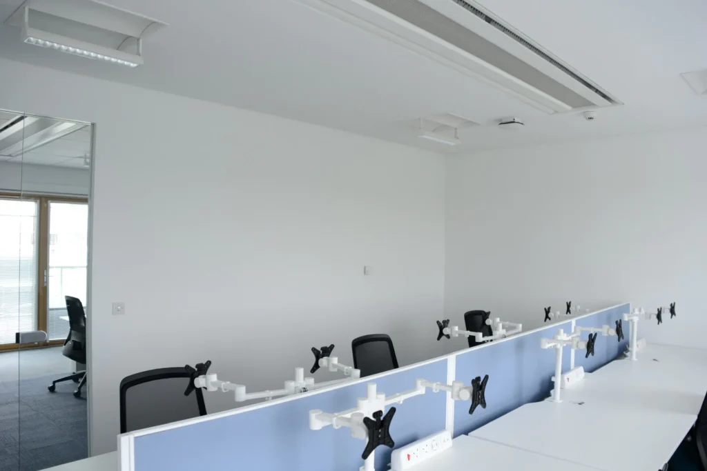 Empty office space with several desks and chairs, blue dividers, and monitor mounts under ceiling lights.