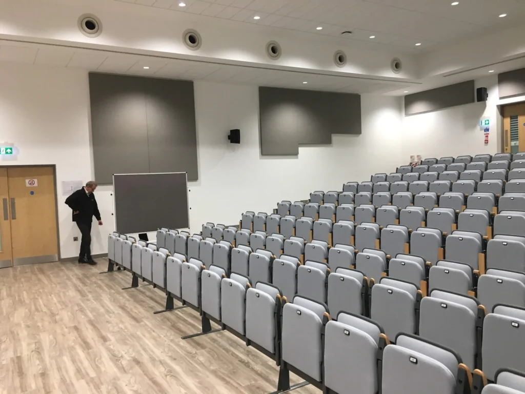 An empty lecture hall with rows of gray seats and a man adjusting a chalkboard.