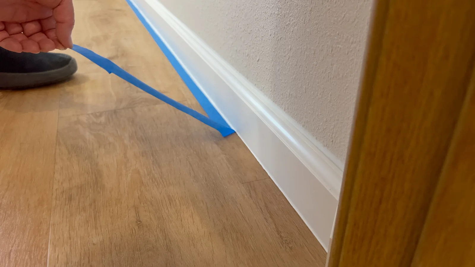 Blue masking tape being pressed along a skirting board over a wooden floor