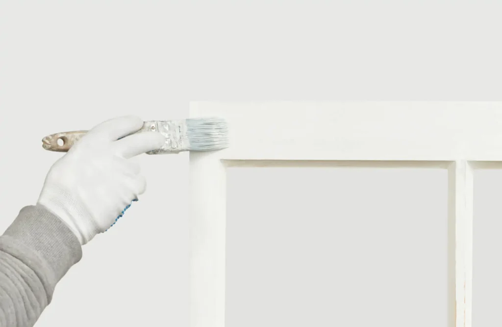 A gloved hand uses a brush to paint a wooden surface white.